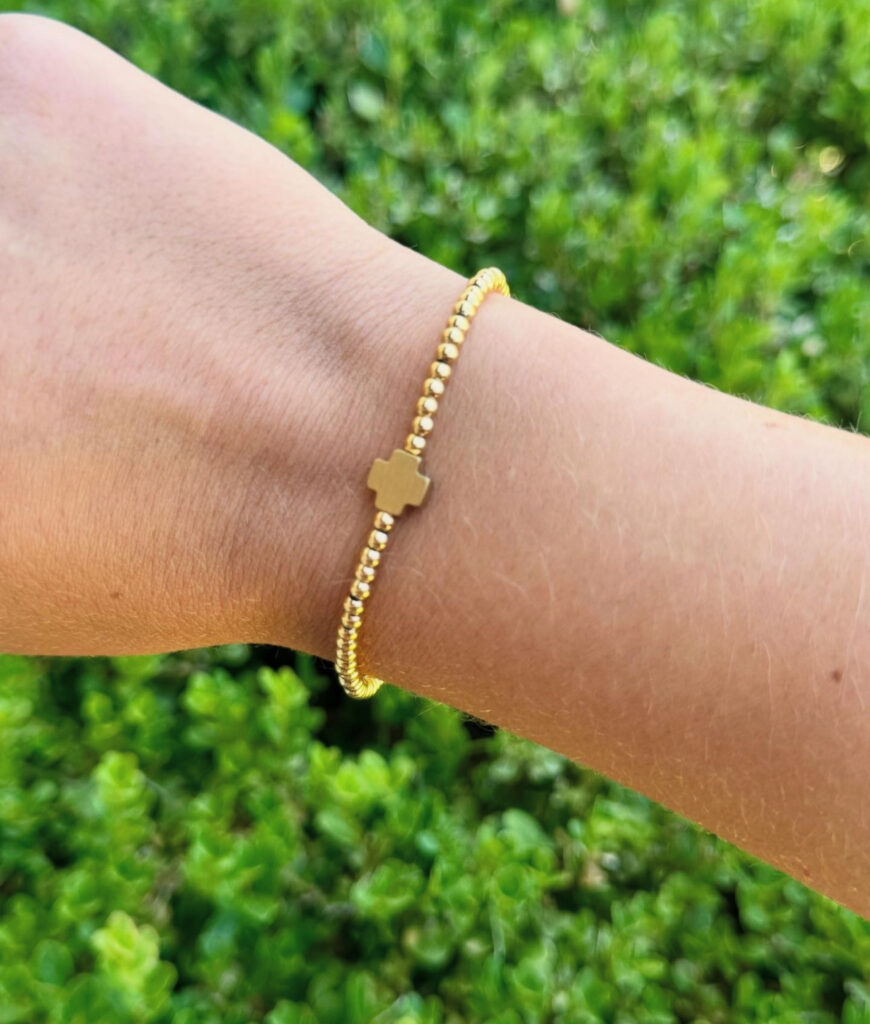 Gold beaded stretch bracelet with a small cross charm, worn on a wrist against a green leafy background.