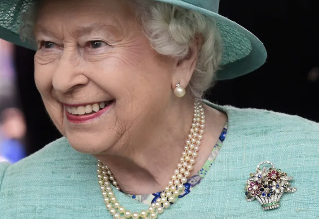 Queen Elizabeth II: Queen Elizabeth II smiles in a turquoise hat and matching jacket, wearing pearl jewelry and a colorful jeweled flower-basket brooch.
