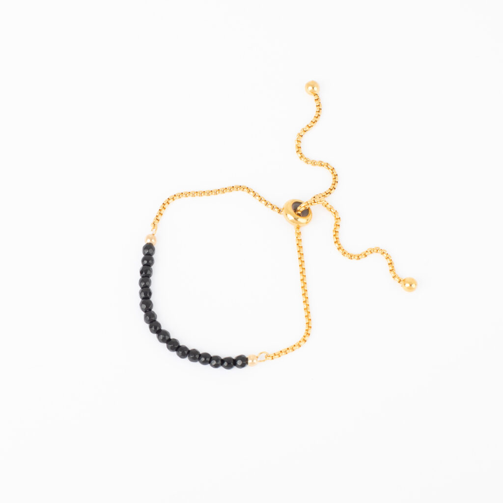 Close-up of a hand wearing two black beaded bracelets, including one with a small black charm and pearl accent, paired with stacked gold rings against a light denim background.