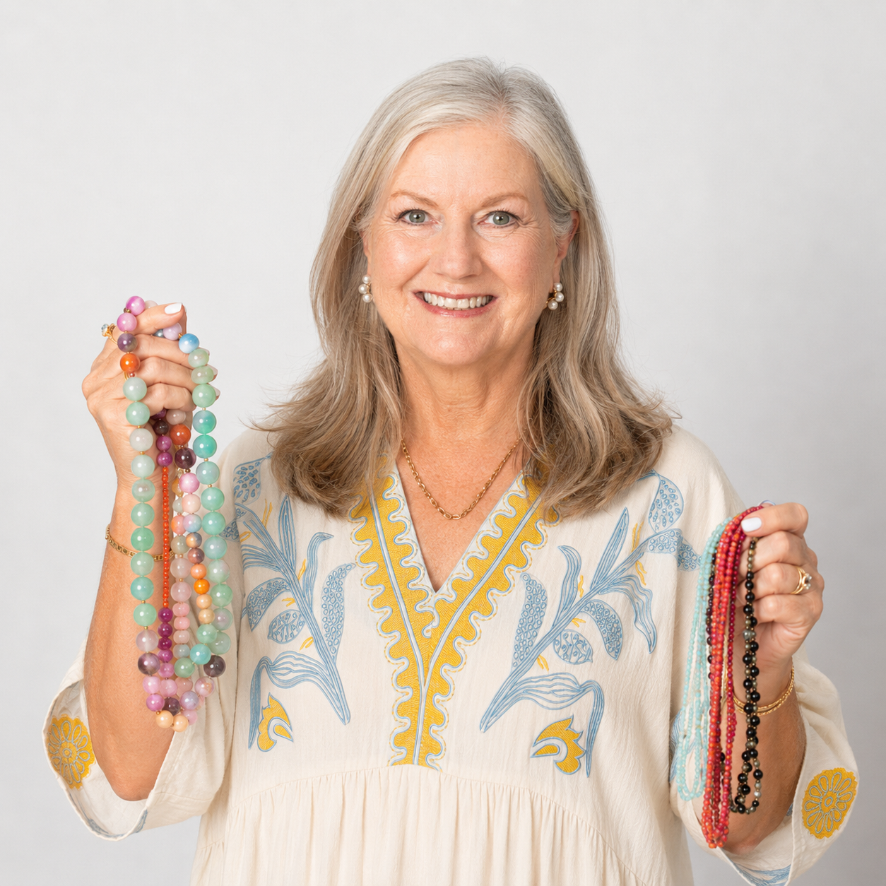 Smiling woman with shoulder-length silver hair wearing a cream embroidered dress with blue and yellow details. She holds colorful beaded statement necklaces in one hand and delicate seed bead necklaces in the other against a soft gray studio background. The portrait highlights vibrant handmade jewelry in a clean, professional setting.
