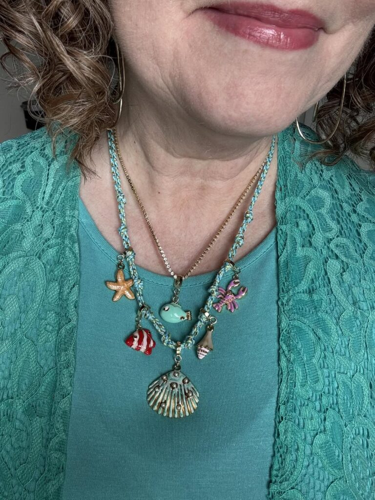 Layered coastal charm necklaces with fish, shell, starfish, and crab charms worn with a teal top.