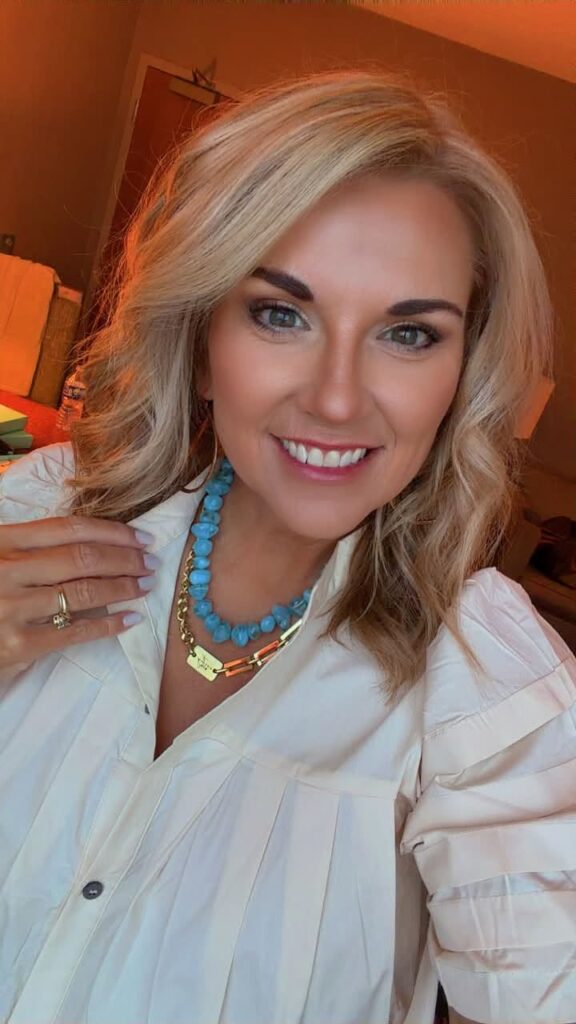 Close-up of a woman wearing a chunky light blue beaded necklace layered with a gold paperclip chain necklace over a white blouse. The soft blue beads add a fresh pop of color while showing how oversized beaded necklaces can be layered with everyday gold jewelry for a modern Fall 2026 look.