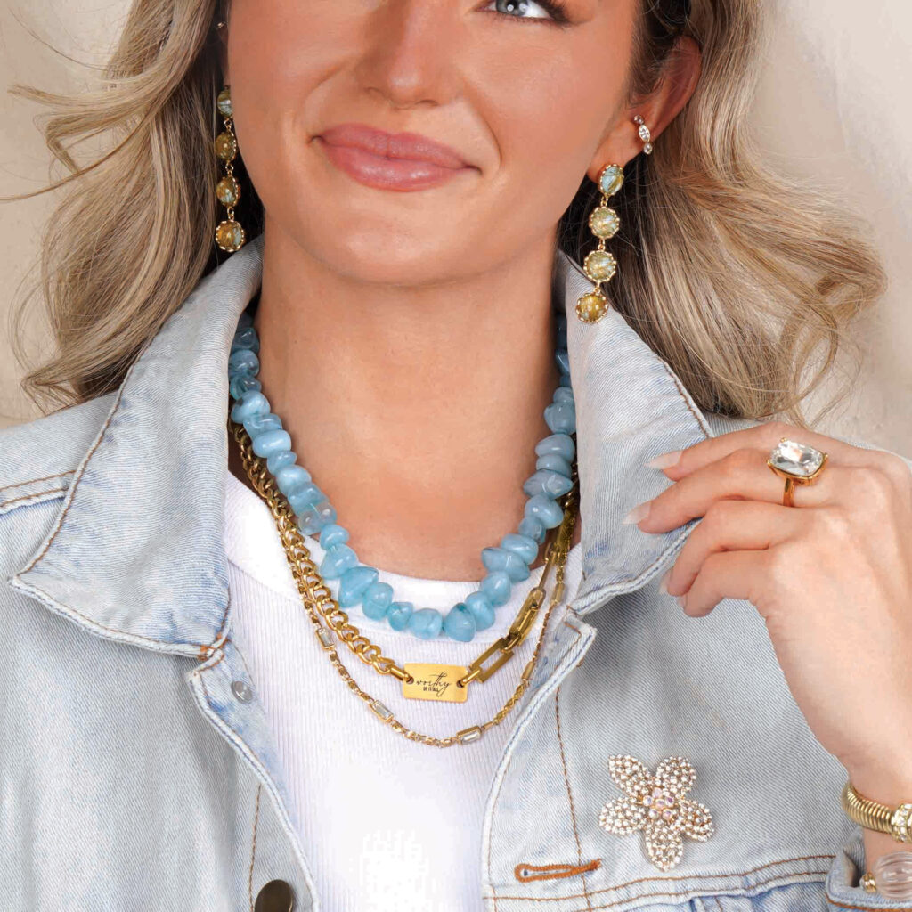 Woman wearing the Mera aqua beaded necklace layered with gold chains, a white top, and a light denim jacket.