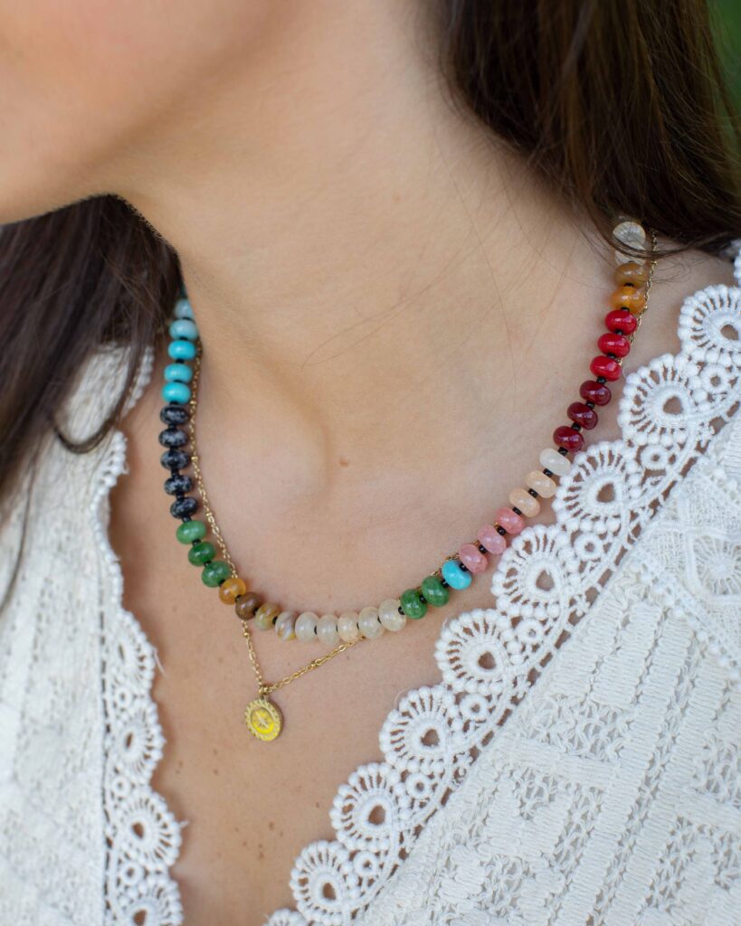 Close-up of a woman wearing the Gertie colorful natural bead necklace with a gold pendant necklace and a white lace blouse.