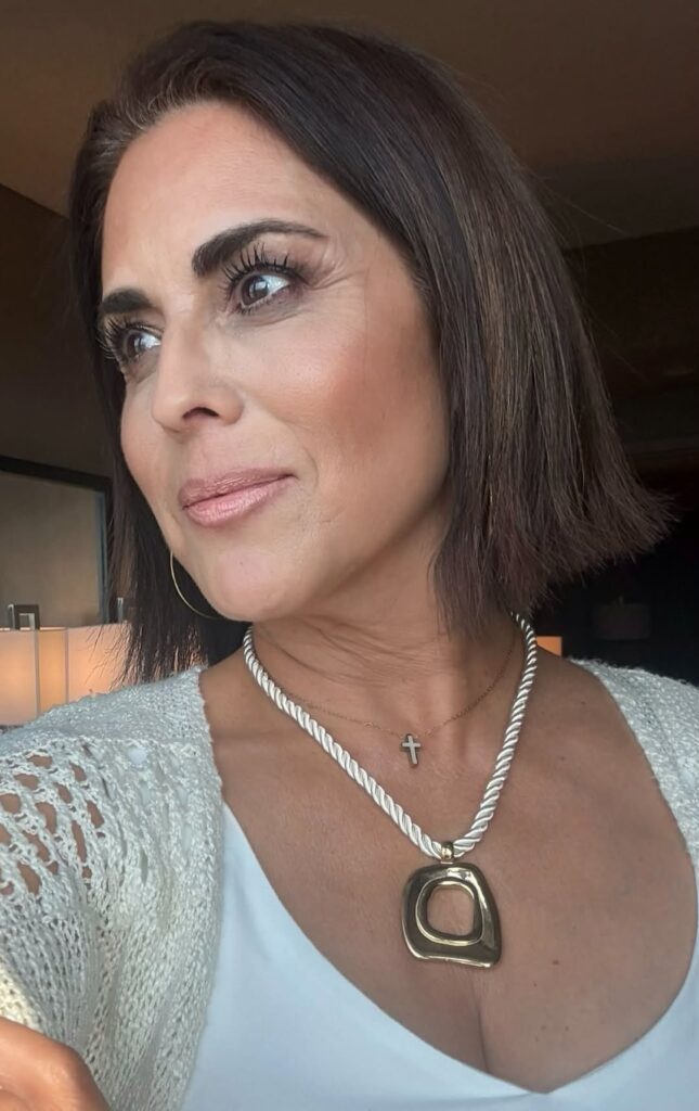 Woman wearing the gold Louisa necklace with a small cross necklace, white scoop-neck top, and cream cardigan.