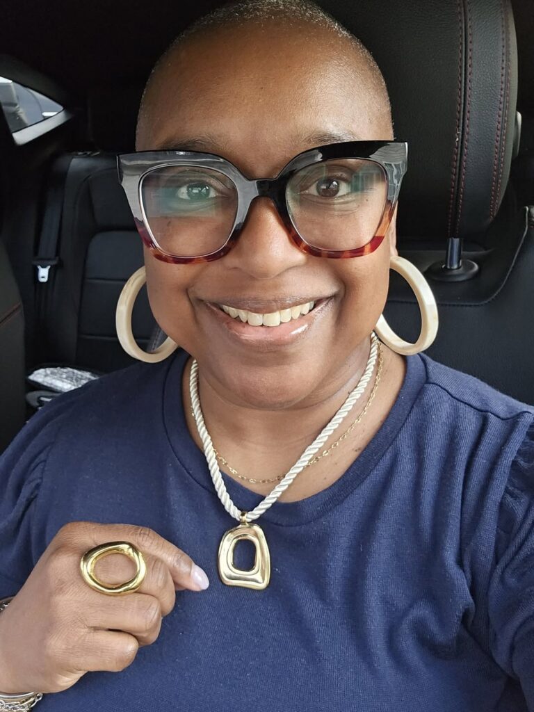 Smiling woman in a navy top wearing the gold Louisa necklace layered with a delicate chain and paired with a bold gold ring.