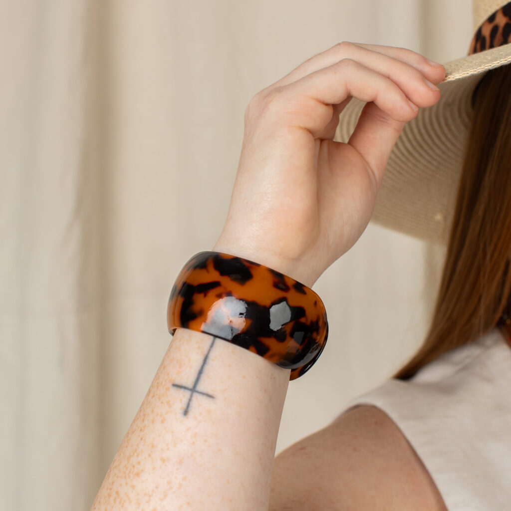 Wide tortoise resin cuff with an amber and brown pattern worn with a neutral summer outfit and straw hat.