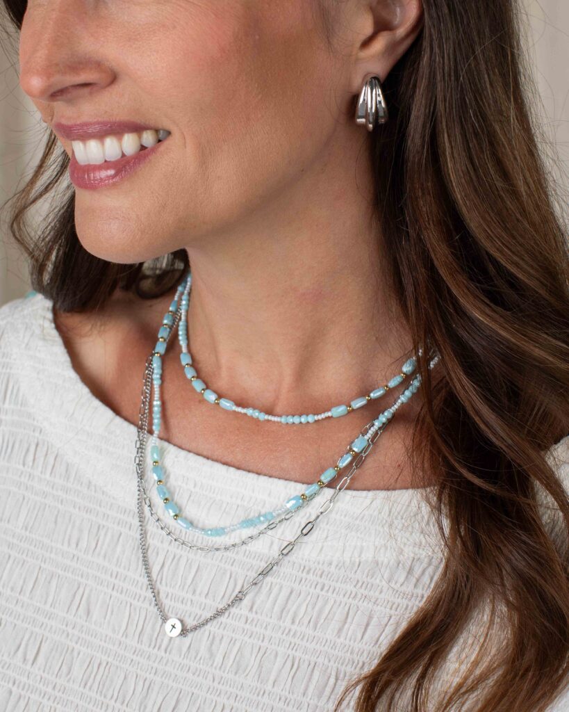 Woman wearing silver James earrings with layered blue and silver necklaces and a white top.