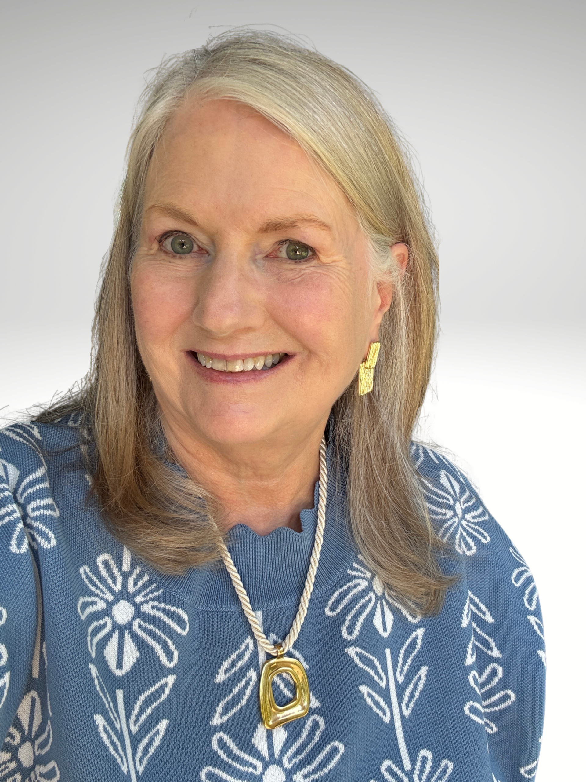 Audrey Peacock smiles in a blue floral sweater, wearing gold drop earrings and a gold pendant on a cream cord necklace.