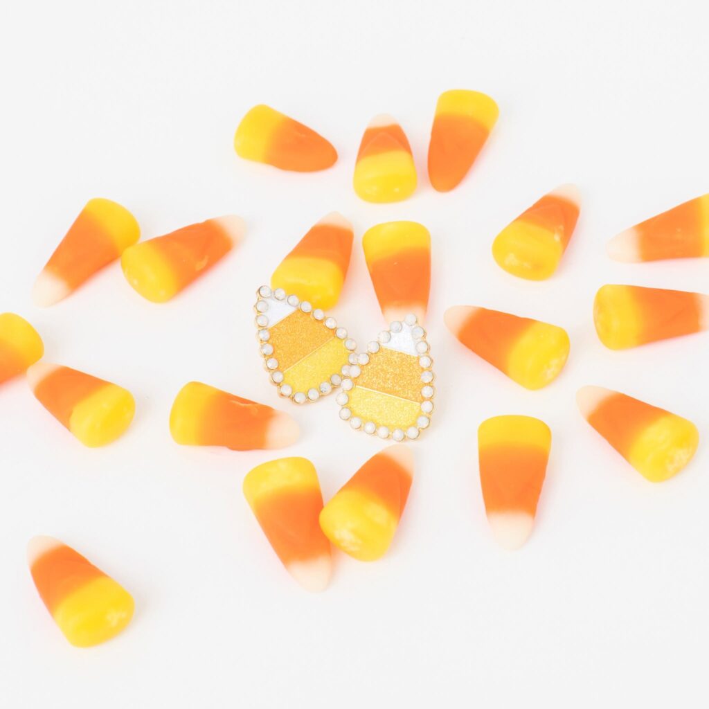 Sparkling candy corn earrings surrounded by orange, yellow, and white candy corn on a white background.