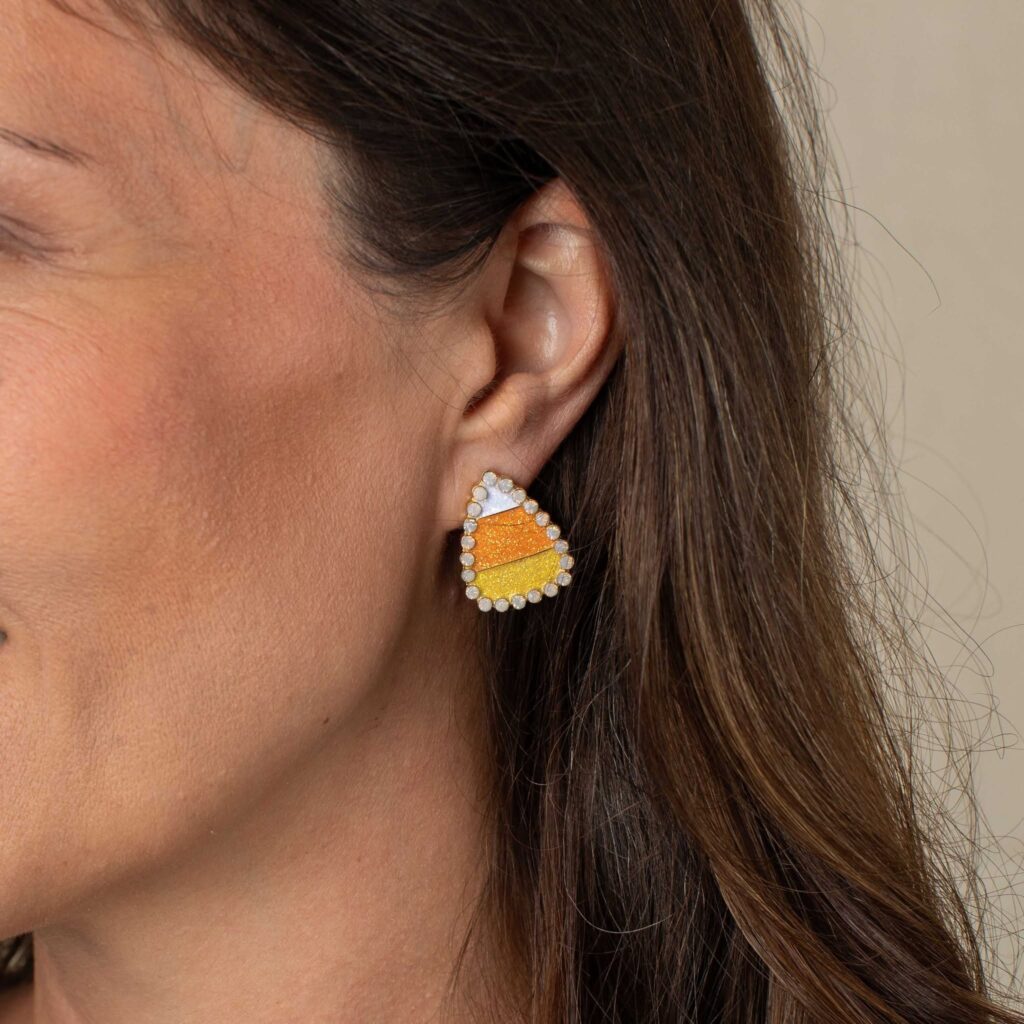 Woman wearing an orange, yellow, and white candy corn earring bordered with small sparkling stones.