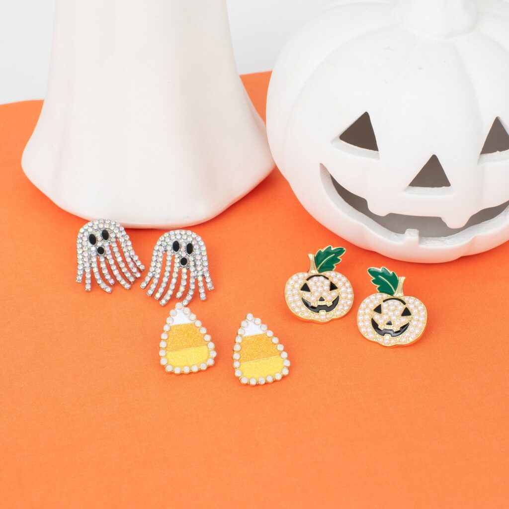 Sparkling ghost, candy corn, and pearl jack-o’-lantern earrings arranged with white Halloween decorations on an orange background.