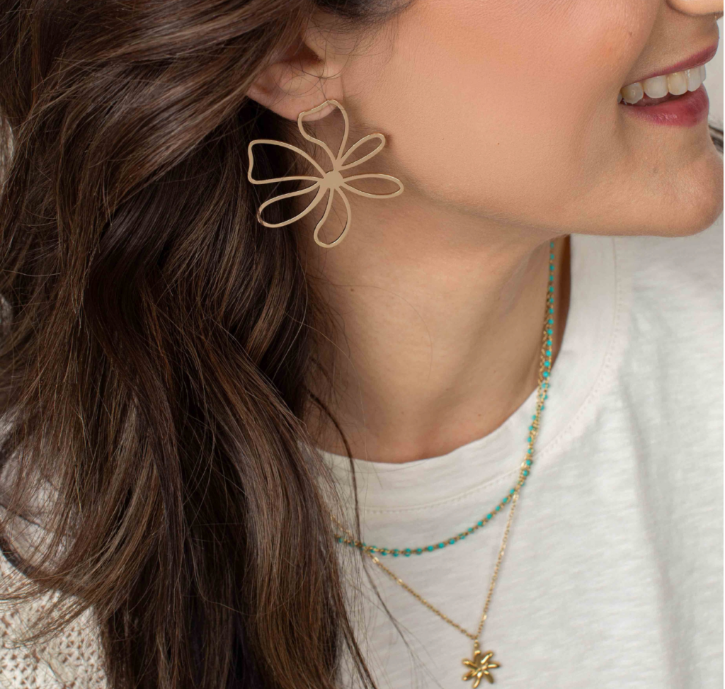 Close-up of a woman wearing a gold Talia flower earring with a white top and layered turquoise necklaces.