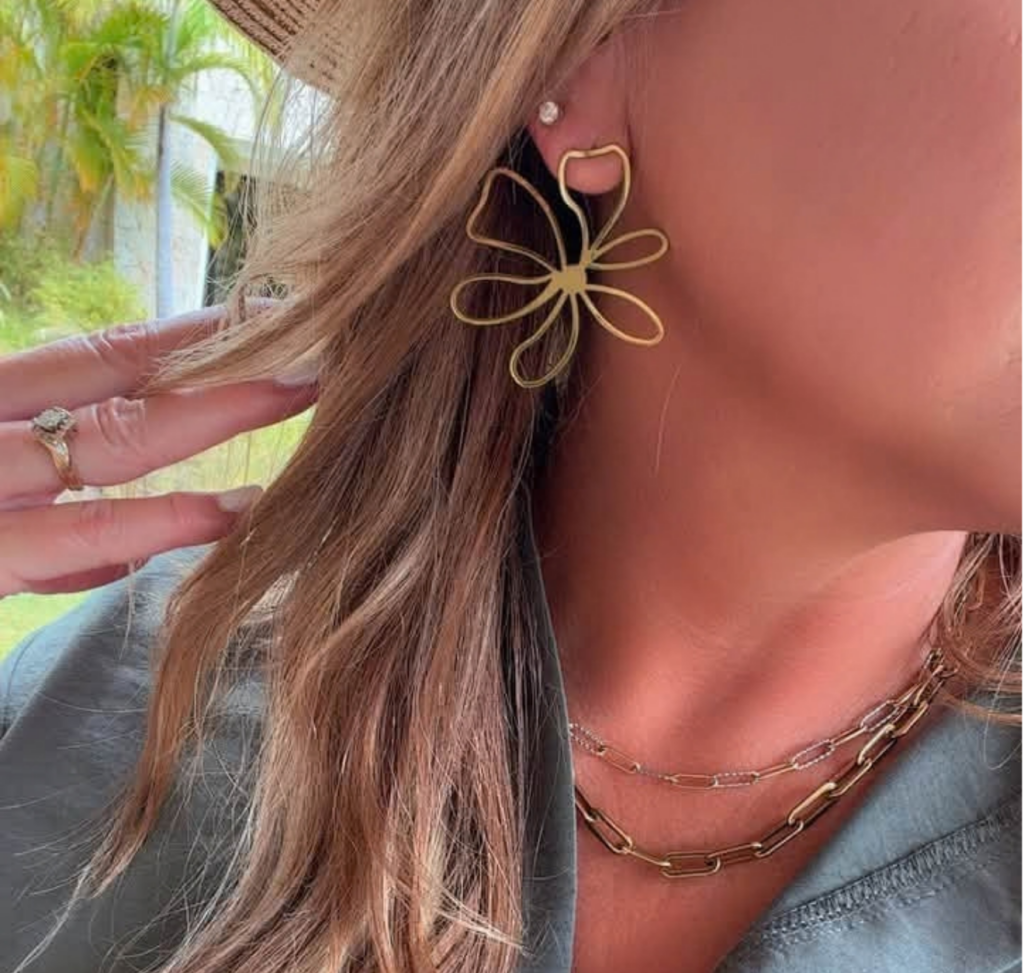 Close-up of a gold Talia flower earring styled with layered gold necklaces and a gray-green top