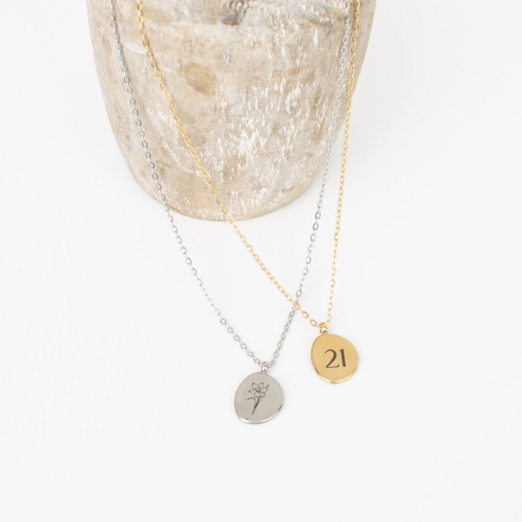 Leya Necklaces in silver and gold with round pendants, one engraved with a flower and the other with “21,” displayed on a wooden stand.