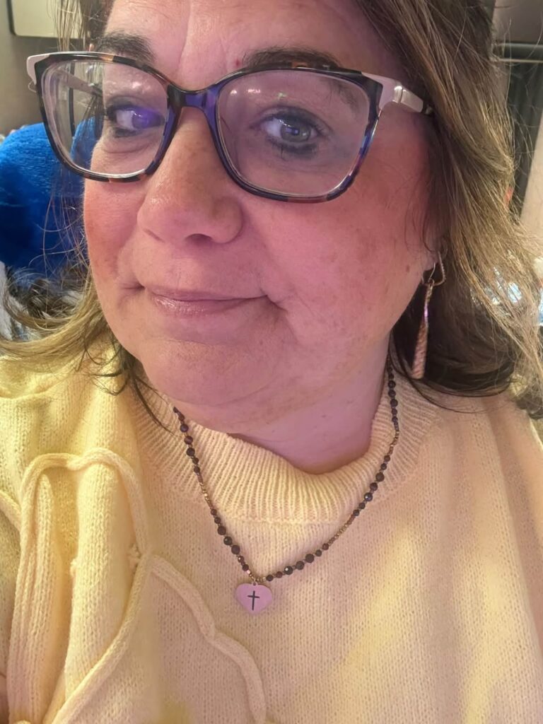 Angela beaded heart necklace personalized with a cross, worn over a pale yellow sweater