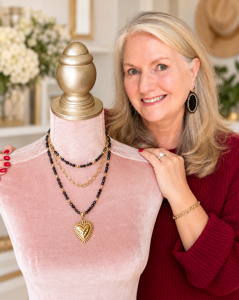 Audrey Peacock of Peacock’s Pretties smiling beside a pink velvet jewelry display featuring layered black and gold necklaces with a gold heart pendant.