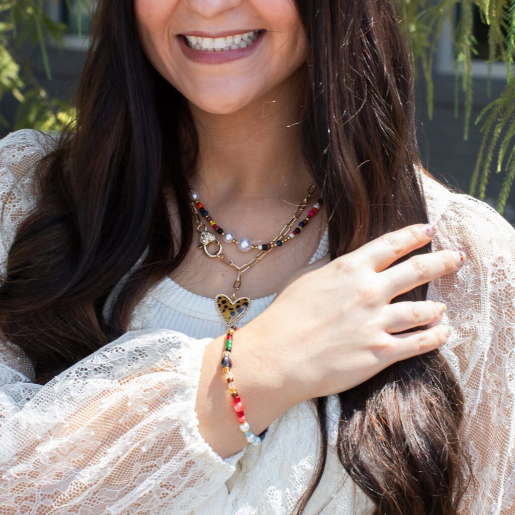 Woman in a white lace blouse wearing jBloom October Bloom Box jewelry: a colorful beaded necklace layered with a gold-tone leopard-print heart necklace and a coordinating multicolored beaded bracelet.