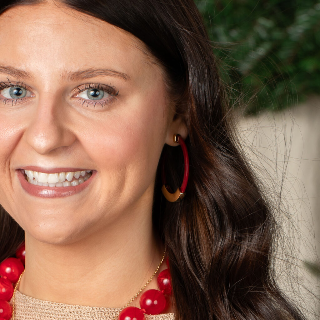 The woman wearing Crimson with a red bead necklace. Woman wearing crimson red hoop earrings with gold accents, a cream knit top, and a red bead necklace.