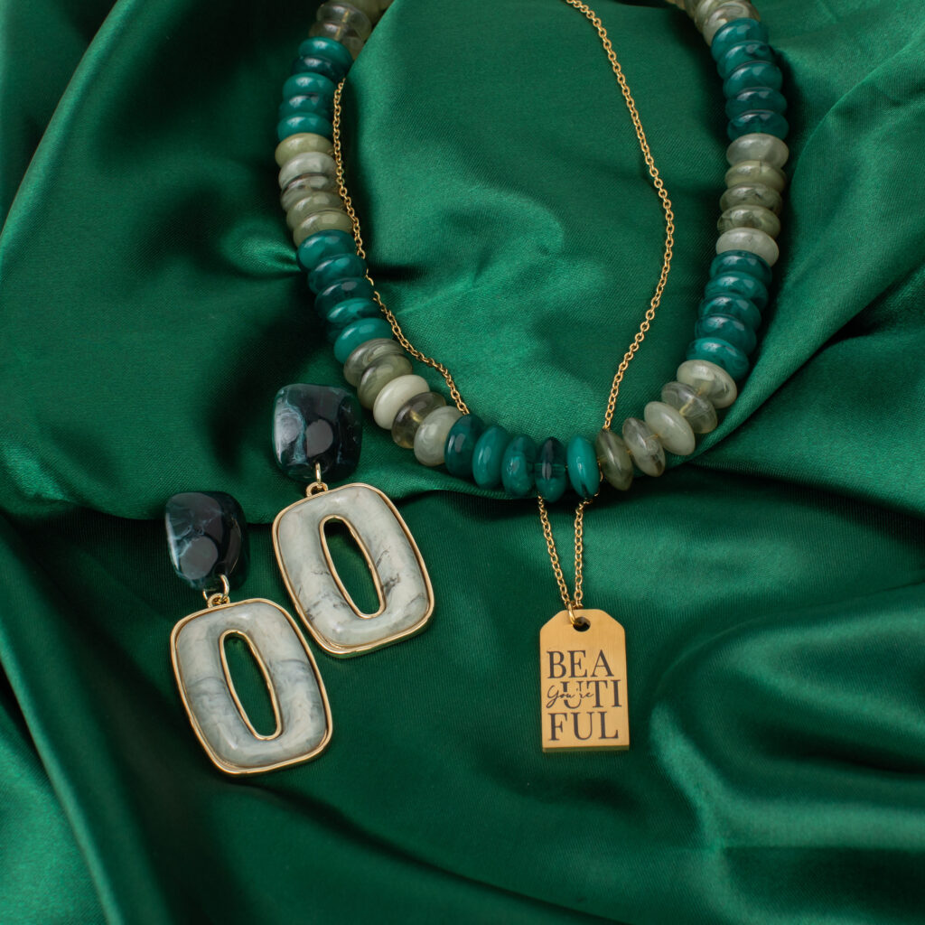 Cypress beaded necklace styled on emerald fabric with a separate gold pendant necklace and rectangular drop earrings.