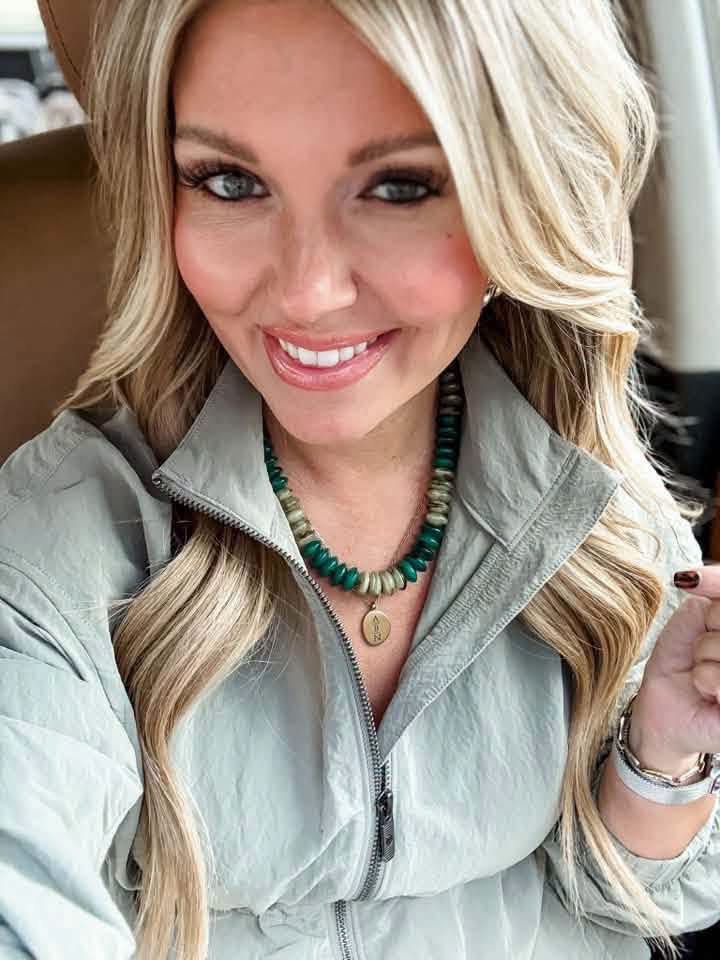 Woman wearing the Cypress green beaded necklace with a pale sage zip-neck top and a separate gold pendant necklace.