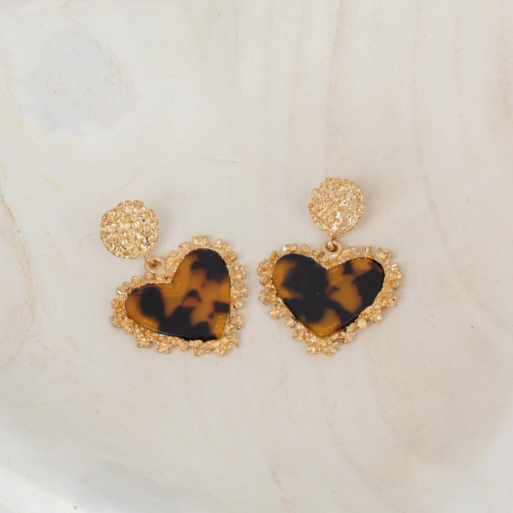 Pair of amber-and-black tortoiseshell-pattern heart earrings with decorative gold-tone borders and textured round studs, displayed on a light stone surface.